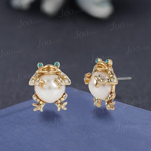 May include: A pair of gold-coloured frog-shaped stud earrings. Each frog has a large, round, white pearl body, with iridescent accents on the legs and arms. The eyes are small, round, and blue. The earrings are set against a blue background.