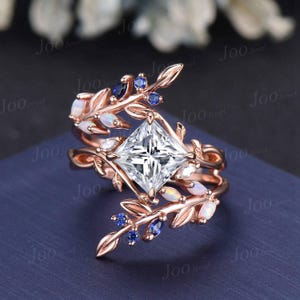 May include: A rose gold engagement ring with a square-cut diamond center stone. The ring features a delicate floral design with opal and sapphire accents.