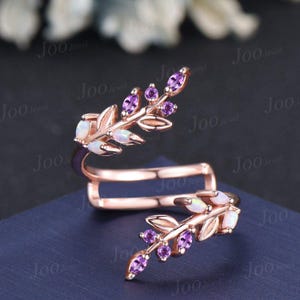 May include: Rose gold ring with a leaf design. The ring features opal and purple amethyst gemstones. The ring is on a blue surface. The ring is a jewelry item.