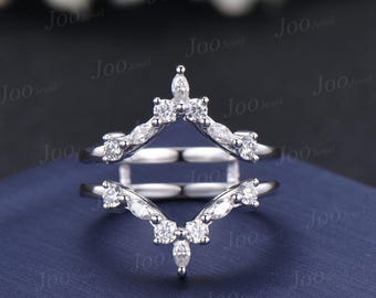 May include: A silver ring guard with a V-shaped design, adorned with multiple small, round, and marquise-cut clear gemstones. The ring guard is designed to sit alongside another ring. The background is a dark blue.