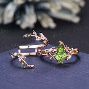 May include: A set of two rose gold rings with a delicate leaf design. The larger ring features a square-shaped green gemstone surrounded by smaller diamonds. The smaller ring is a simple band with a leaf design.