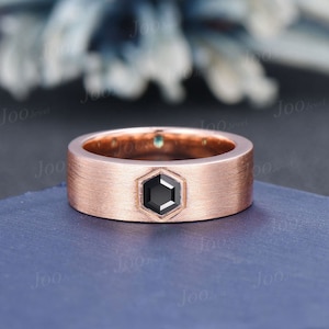 May include: A rose gold ring with a brushed finish. The ring features a hexagonal black gemstone at the center. The ring is set on a blue surface.