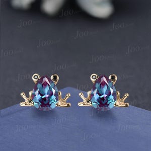 May include: A pair of gold-coloured frog-shaped earrings. Each earring has a pear-shaped, multi-coloured gemstone in teal and purple. The frog's eyes are black, and the legs are outstretched.