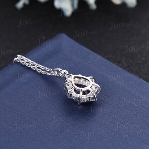 May include: A silver pendant necklace with a halo design. The pendant features a central dark stone surrounded by smaller, clear stones. The necklace chain is delicate and silver. The pendant rests on a blue surface.