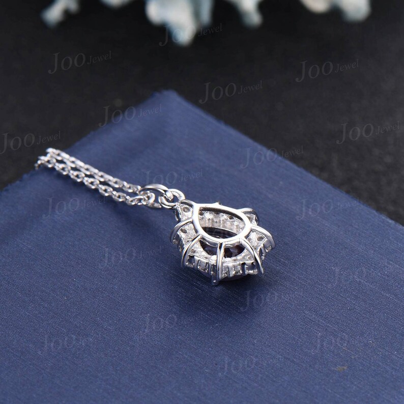 May include: A silver pendant necklace with a halo design. The pendant features a central dark stone surrounded by smaller, clear stones. The necklace chain is delicate and silver. The pendant rests on a blue surface.