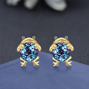 May include: A pair of gold-coloured frog-shaped earrings. Each earring has a large, round, blue and purple gemstone body with small turquoise eyes. The earrings are set against a blue and grey background.