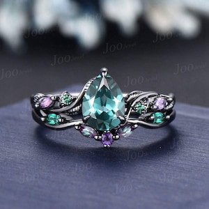 May include: A silver-toned ring with a large, pear-shaped teal gemstone as the focal point. The band features intricate leaf and vine detailing, with smaller green, purple, and pink gemstones. The ring is set against a dark blue background.