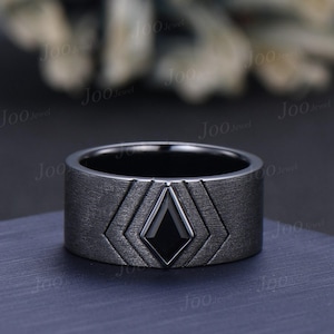 Op de afbeelding: Een brede, donkergrijze bandring met een getextureerde, geborstelde metalen afwerking. Het midden van de ring toont een geometrische, ruitvormige zwarte inleg omringd door gegraveerde, concentrische chevronlijnen die naar het midden wijzen.