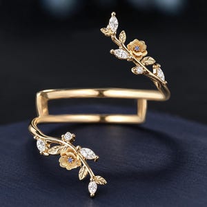 May include: A gold-toned open ring featuring two branching ends, each adorned with small sculpted flowers and marquise-cut clear stones. The band has a rectangular shape, and the floral motifs include tiny blue centers within the petals.