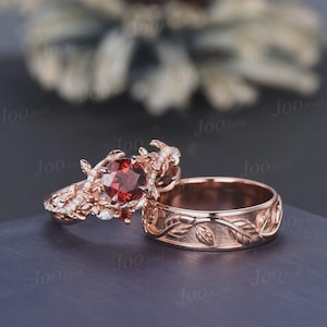 May include: A rose gold ring set featuring a round, deep red garnet center stone surrounded by intricate leaf and vine metalwork with small white accent stones, paired with a matching rose gold band engraved with a continuous leaf pattern.