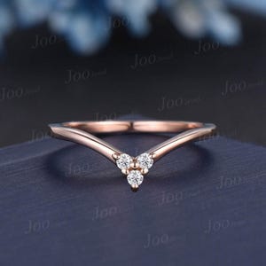 May include: A rose gold V-shaped chevron ring featuring three small, round, clear, prong-set stones arranged in a triangular cluster at the center point of the band. The metal has a smooth, polished finish throughout the entire ring structure.