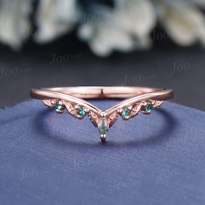 Könnte beinhalten: Ein zarter Ring aus Roségold mit einem V-förmigen Design und kleinen grünen Edelsteinen. Der Ring ist mit einem größeren grünen Edelstein an der Spitze des V besetzt.