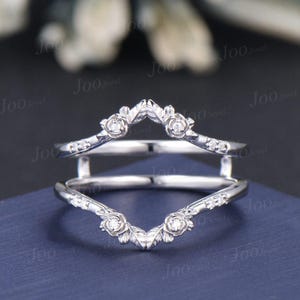 May include: A silver ring guard with a floral design. The ring features small, round gemstones and rose-shaped accents. The guard is designed to sit around another ring, enhancing its appearance. The ring is displayed on a blue surface.