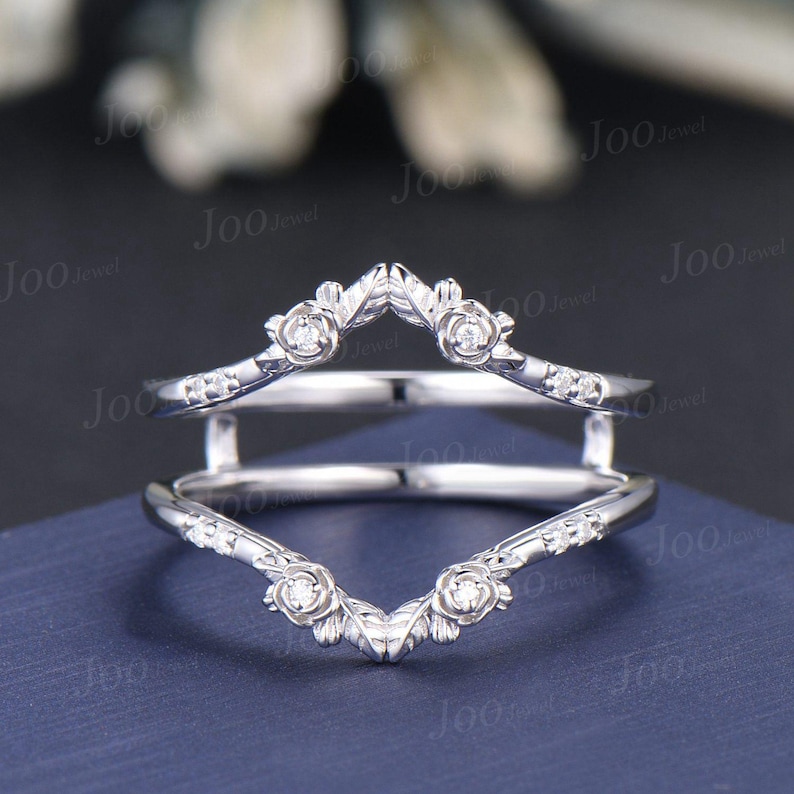 May include: A silver ring guard with a floral design. The ring features small, round gemstones and rose-shaped accents. The guard is designed to sit around another ring, enhancing its appearance. The ring is displayed on a blue surface.