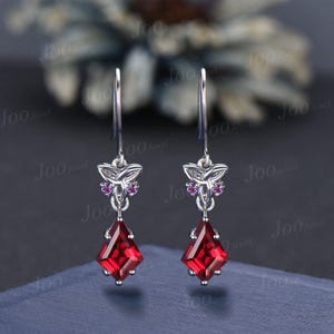May include: A pair of silver dangle earrings featuring a red gemstone and a small butterfly charm with pink accents.