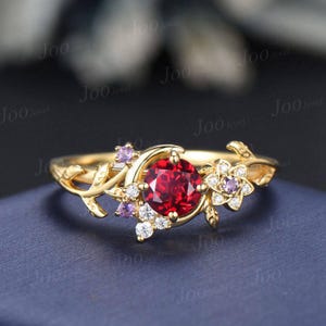 May include: A gold-colored ring featuring a large, round, red gemstone at the center. The ring is adorned with floral and leaf designs, and smaller white and purple gemstones. The ring has a delicate, nature-inspired aesthetic.