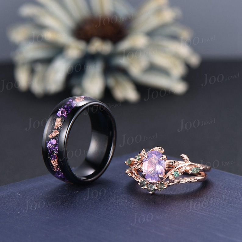 Puede incluir: Dos anillos: una banda negra con motas moradas y doradas, y un anillo de oro rosa con una piedra ovalada de color p&uacute;rpura claro y detalles verdes. Los anillos se exhiben sobre una superficie azul oscuro.