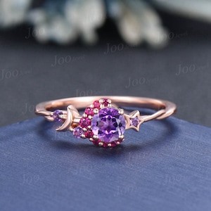 May include: A rose gold ring with a purple amethyst gemstone surrounded by smaller pink gemstones. The ring features a crescent moon and star design.