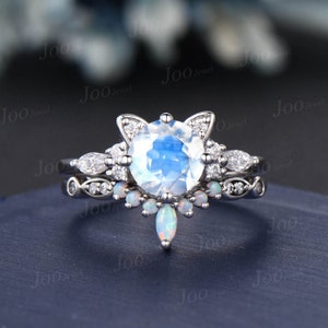 May include: A silver ring set featuring a large, round, iridescent moonstone center stone, accented with small diamonds and opals. The main ring has a cat ear design. The set includes a matching band with small stones.