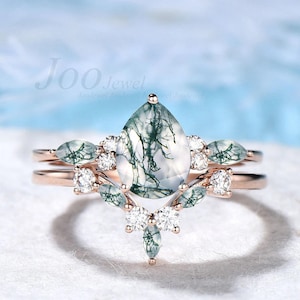 May include: A rose gold ring set featuring a pear-shaped moss agate center stone with green inclusions. The ring is accented with small white diamonds and green marquise-cut stones. The band is delicate and the overall design is elegant.