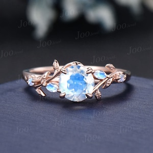 Könnte beinhalten: Roségoldener Ring mit einem großen, runden, irisierenden Mondstein. Der Ring ist mit zarten Blättern und kleinen Mondstein-Akzenten verziert. Der Ring liegt auf einer dunkelblauen Oberfläche.