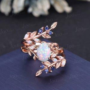May include: A rose gold ring with a white opal gemstone surrounded by delicate leaves and blue sapphires. The ring is designed with a floral motif and features a unique, intertwined band.