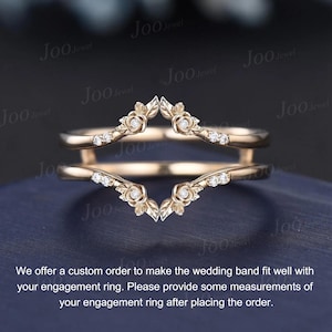 May include: A gold-colored wedding band with a floral design and small, sparkling stones. The band has a unique, curved shape, designed to fit around an engagement ring. The band is shown on a dark blue surface.