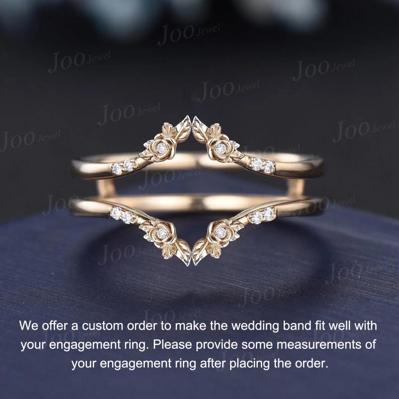 May include: A gold-colored wedding band with a floral design and small, sparkling stones. The band has a unique, curved shape, designed to fit around an engagement ring. The band is shown on a dark blue surface.