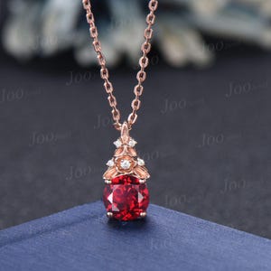 May include: A rose gold necklace with a pendant featuring a large, round, red gemstone surrounded by smaller, clear gemstones. The pendant is in a floral design.