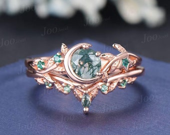 May include: A rose gold ring set featuring a round, moss agate gemstone, a crescent moon, and star accents. The band is adorned with leaf and small green gemstone details. The ring set is displayed on a blue surface.
