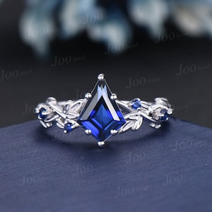 May include: A silver ring with a large, kite-shaped, deep blue gemstone as the centerpiece. The band features a delicate leaf and small blue stone design. The ring is set against a dark blue background.