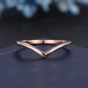 May include: A rose gold V-shaped ring. The ring has a smooth, polished finish and a minimalist design. The band curves to a point at the center, creating a modern and elegant look. The ring is set against a dark blue background.