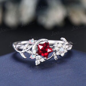 May include: A silver-toned ring featuring a central star-shaped deep red gemstone, accented by small round opals and clear white stones. The band is designed with a vine-like motif, incorporating delicate leaf details and additional small clear stones.