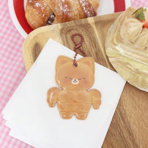 May include: A tan-colored, cartoon-style keychain in the shape of a muscular cat. The keychain has a small, brown chain and a smiling face. The cat's body is detailed with defined muscles. The keychain is on a white napkin.