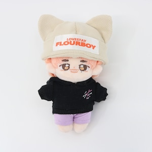 May include: A plush doll wearing a beige beanie with the text "LOVESTAY FLOURBOY" in orange, a black hoodie, and purple shorts. The doll has peach-colored hair and embroidered facial features. The hoodie has a white and pink graphic.