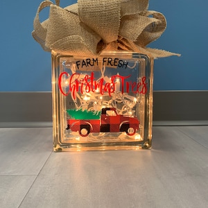 May include: A glass block with a burlap bow on top. The block has a red truck with a Christmas tree on the back. The text on the block reads "Farm Fresh Christmas Trees Pine Spruce Fir Cedar".