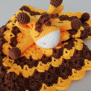 May include: A crocheted baby blanket with a yellow and brown pattern. A crocheted giraffe toy is attached to the blanket.