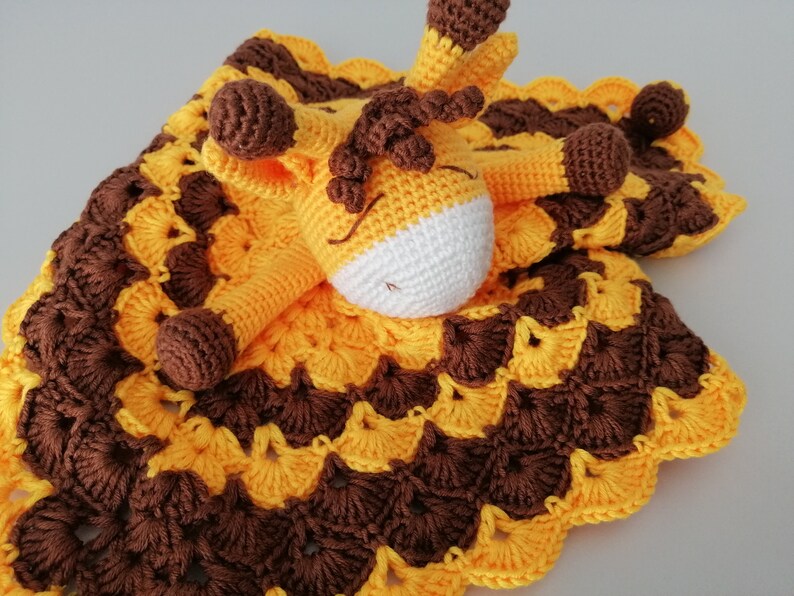 May include: A crocheted baby blanket with a yellow and brown pattern. A crocheted giraffe toy is attached to the blanket.
