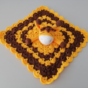 May include: A crocheted baby blanket with a yellow and brown pattern. A small crocheted giraffe is attached to the centre of the blanket.