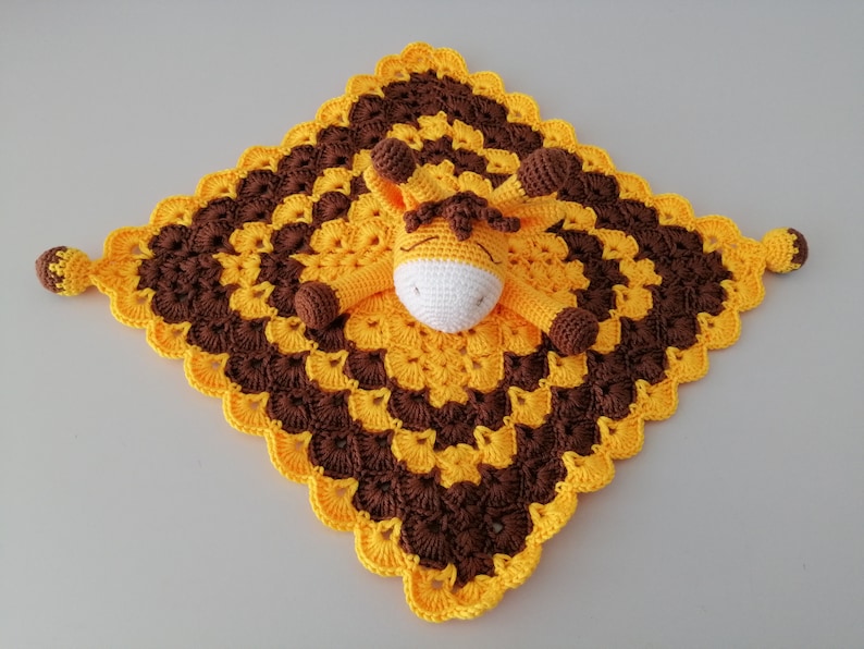May include: A crocheted baby blanket with a yellow and brown pattern. A small crocheted giraffe is attached to the centre of the blanket.