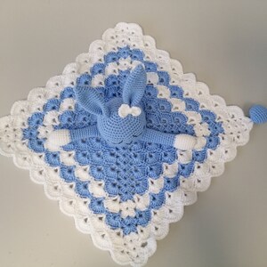 May include: A blue and white crocheted baby blanket with a blue crocheted bunny attached. The blanket has a scalloped edge and the bunny has a white bow.