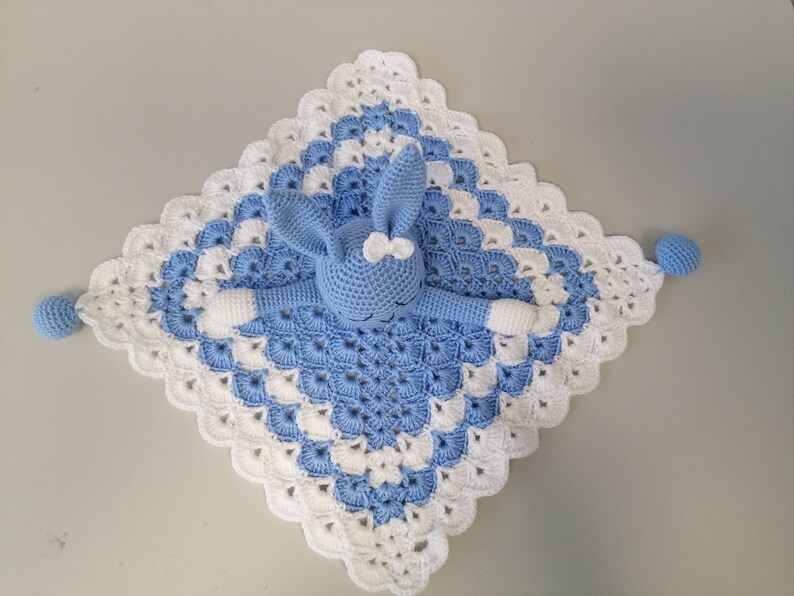 May include: A blue and white crocheted baby blanket with a blue crocheted bunny attached. The blanket has a scalloped edge and the bunny has a white bow.