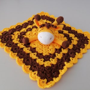 May include: A yellow and brown crocheted baby blanket with a crocheted giraffe attached. The giraffe has a white face and brown eyes.