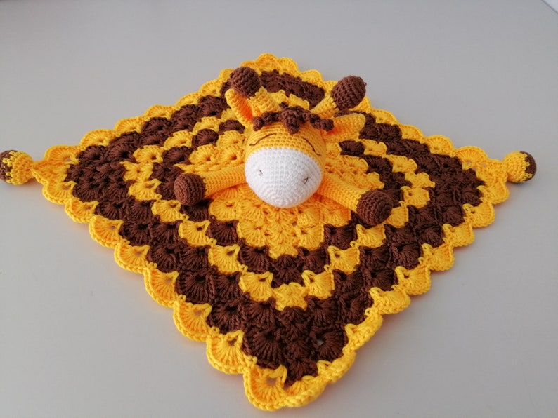 May include: A yellow and brown crocheted baby blanket with a crocheted giraffe attached. The giraffe has a white face and brown eyes.