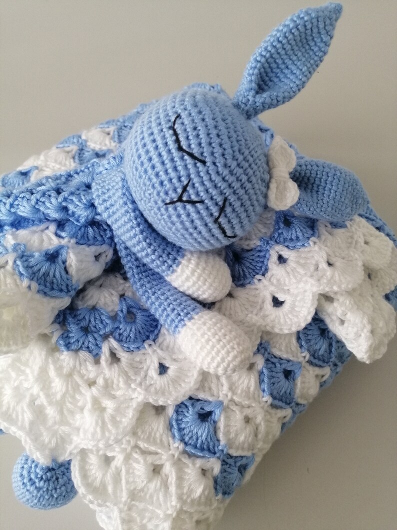 May include: A blue crocheted bunny with a white bow, lying on a blue and white crocheted blanket.