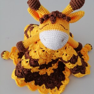 May include: A crocheted giraffe soft toy with a yellow and brown blanket. The giraffe has brown ears and a white face.