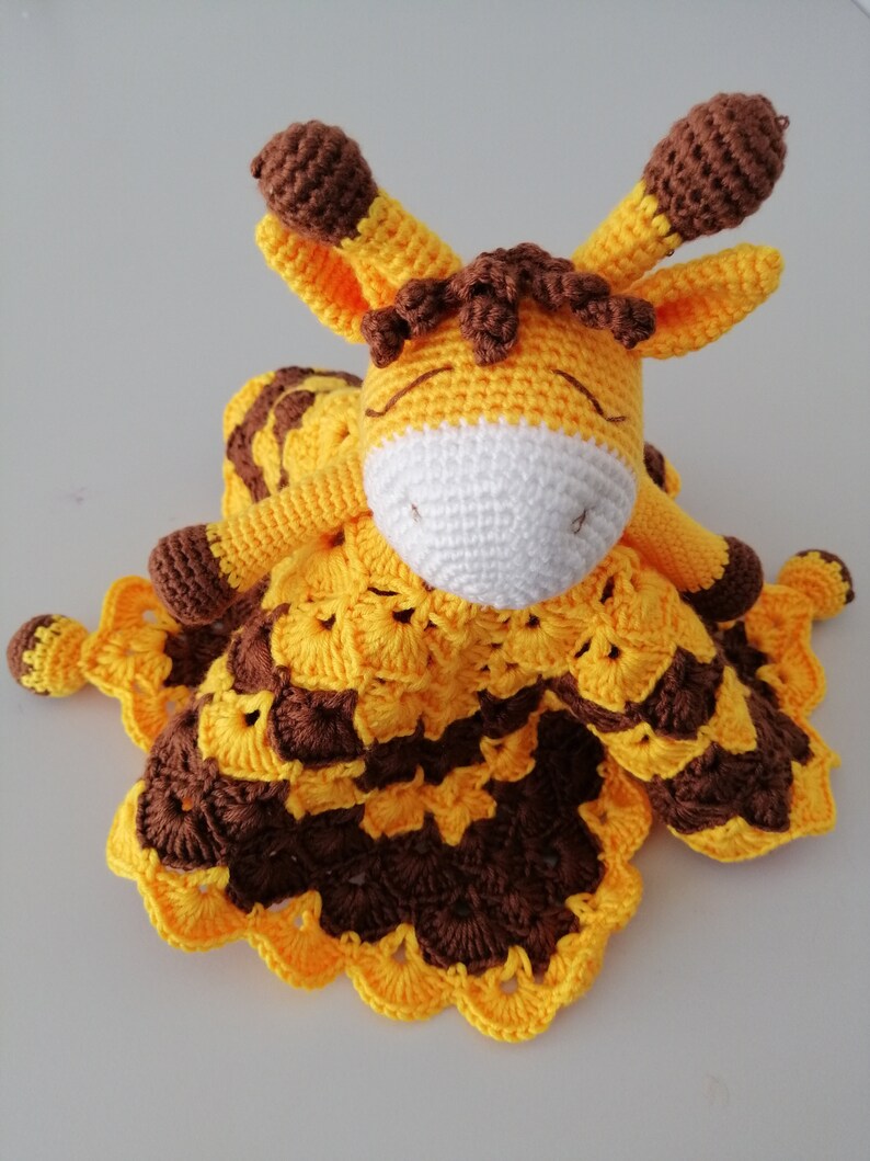 May include: A crocheted giraffe soft toy with a yellow and brown blanket. The giraffe has brown ears and a white face.