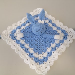 May include: A blue crocheted baby blanket with a white crocheted border. A blue crocheted bunny with white accents is attached to the blanket.