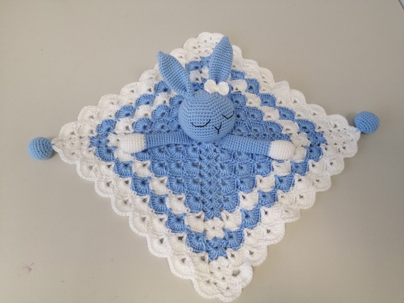May include: A blue crocheted baby blanket with a white crocheted border. A blue crocheted bunny with white accents is attached to the blanket.