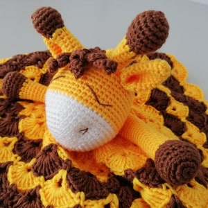 May include: A crocheted giraffe soft toy with brown spots and a yellow and brown blanket. The giraffe is lying on the blanket with its head resting on the blanket.